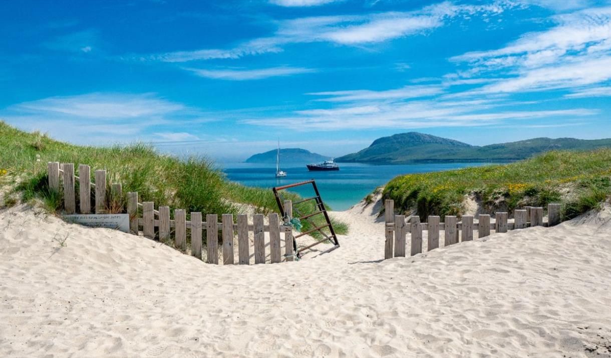 A picturesque view of Vatersay, Isle of Barra, with a wooden fence and gate opening to sandy dunes and the beach beyond.