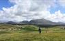 A man walks through green croft fields with scattered houses and mountains in the background.