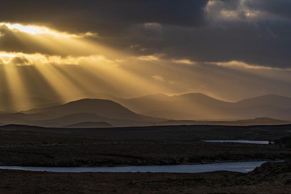 Landscape photo of a sunset over the Lochs area and Lewis and Harris hills. Sunbeams shine onto the landscape through holes in the clouds, create suns