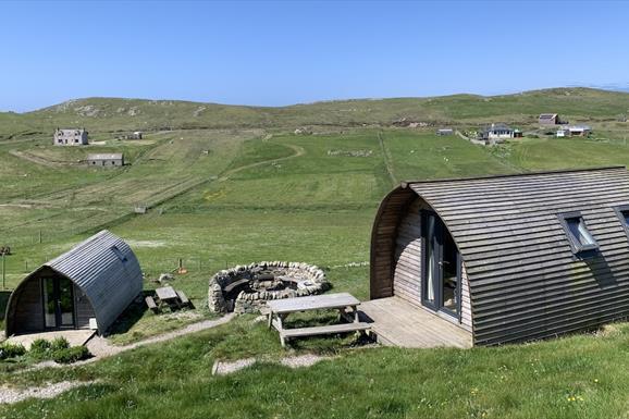 Two curved wooden cabins with picnic tables sit in a grassy meadow with a circular stone fire pit between them.